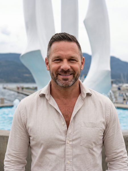 JC Rathwell smiling in a linen shirt in front of the Spirit of Sail sculpture on Kelowna's waterfront