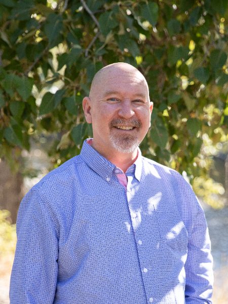 Todd Sanderson smiling in a blue patterned button-up shirt, outdoors in front of a leafy tree