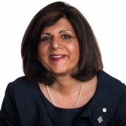 Mohini Singh smiling in a navy jacket and pearl necklace against a white background