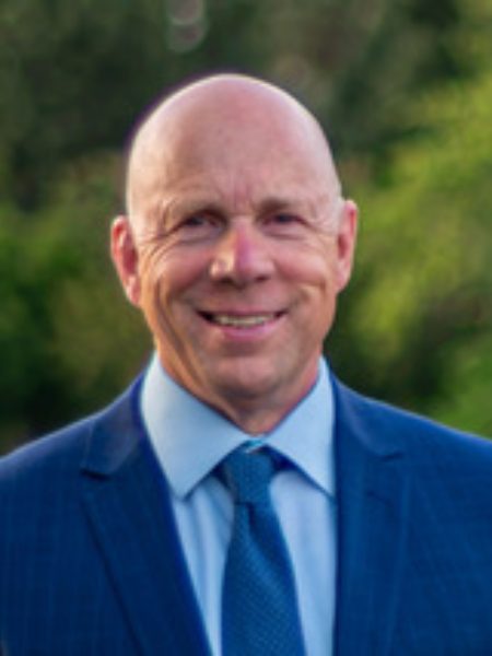 Ron Cannan smiling in a navy suit and blue tie, outdoors in front of green trees