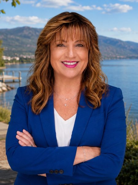 Julia Fraser smiling with arms crossed in a blue blazer, with Okanagan Lake and hills behind her
