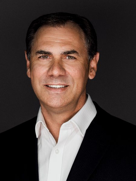 Greg Dahms smiling in a dark blazer and white shirt against a dark background
