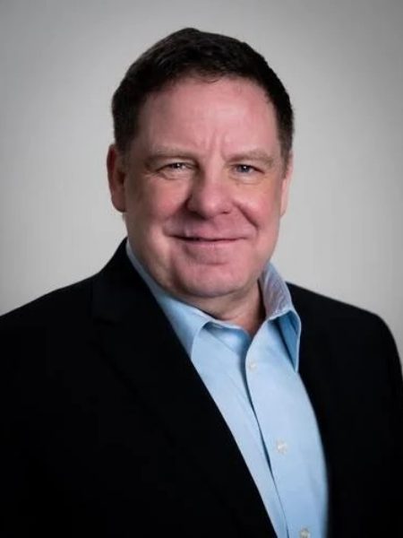 Rick Webber smiling in a dark blazer and light blue shirt against a grey background