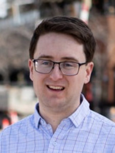 Davis Kyle smiling in glasses and a light blue checked shirt, outdoors on a city street