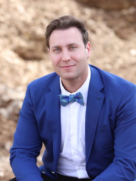Hamid Khajavi in a blue blazer and patterned bow tie, sitting outdoors in front of a rocky slope