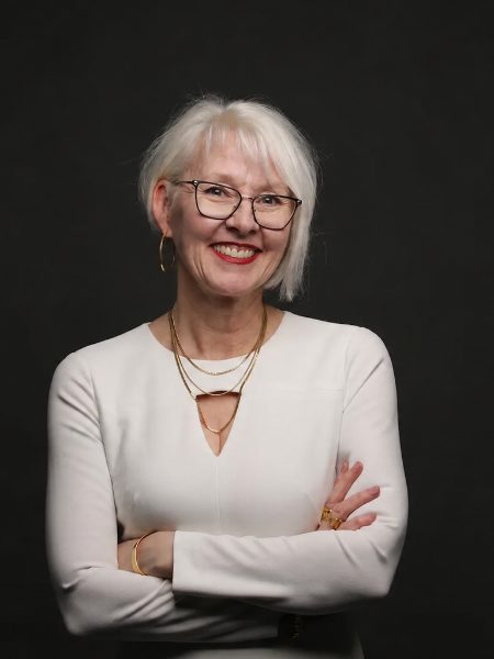Nikki Csek smiling with arms crossed, in a white top and glasses, against a dark background