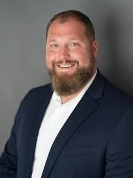 Kevin Kraft smiling, with a full beard, in a navy blazer and white shirt against a grey background