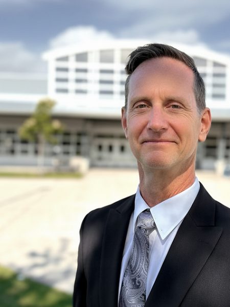 Jim Strachan smiling in a dark suit and grey paisley tie, outside a school building
