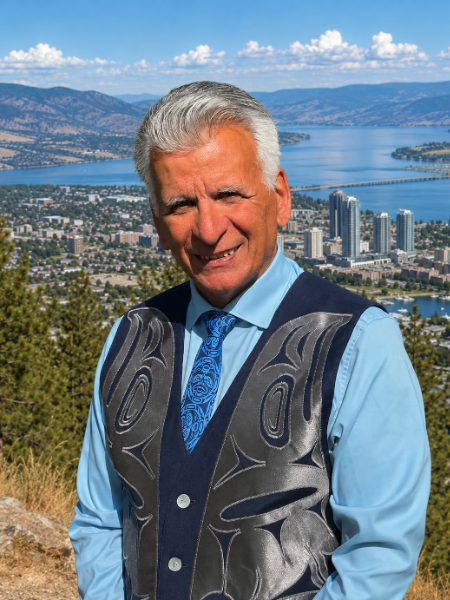 Richard Gauthier smiling in a light blue shirt and a vest with a West Coast Indigenous design, with Kelowna and Okanagan Lake behind him
