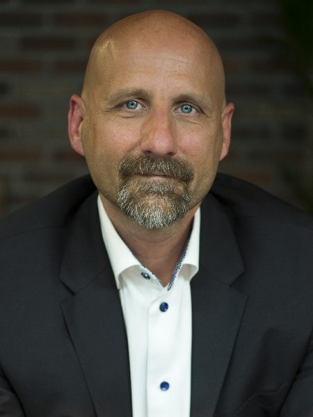Les Bellamy in a dark blazer and white shirt in front of a brick wall
