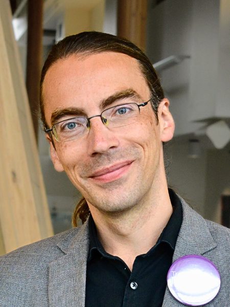 Trevor McAleese smiling in glasses and a grey blazer, with his hair tied back and a campaign button on his lapel
