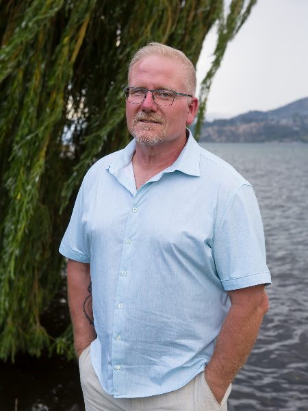 Brian Pinter in glasses and a light blue short-sleeved shirt, standing by the lake under a willow tree