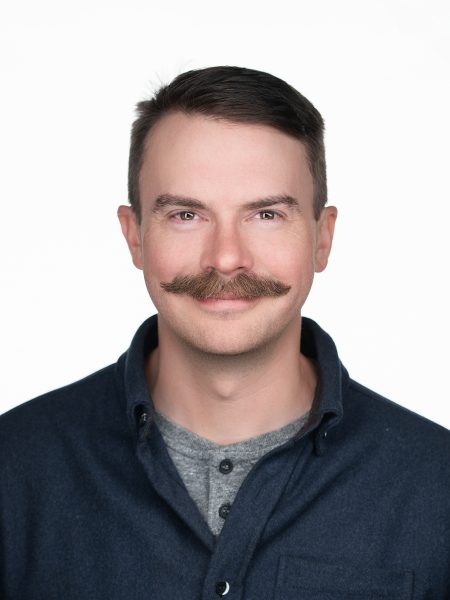 Daniel Fritter with a moustache, in a navy button-up shirt against a white background