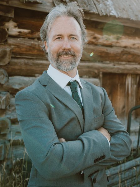 Mike Rose with arms crossed in a grey-green suit and green tie, in front of an old log cabin