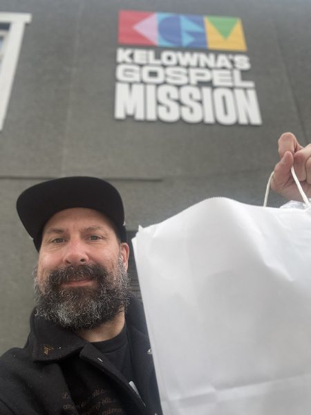 Todd Daniels taking a selfie in a black cap outside Kelowna's Gospel Mission, holding up a white bag