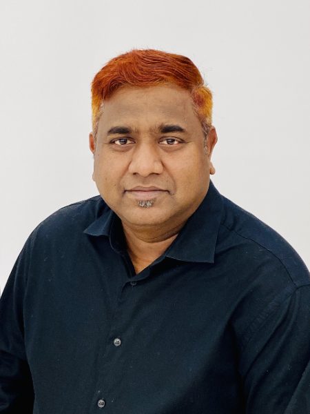 Ashok Kumar Verma with bright orange-dyed hair, in a dark navy button-up shirt against a white wall