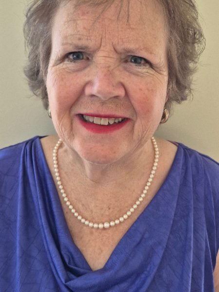 Joyce Brinkerhoff smiling in a blue top and pearl necklace against a cream wall