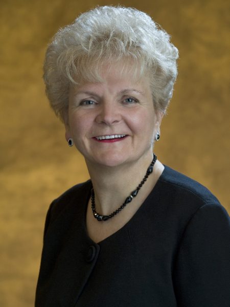 Maxine DeHart smiling in a black top and beaded necklace against a gold backdrop